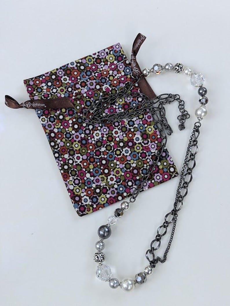 Puede incluir: Un collar plateado con una mezcla de cuentas y eslabones de cadena, junto a una bolsa con estampado floral. El collar presenta cuentas transparentes, grises y perladas. La bolsa tiene una cinta marr&oacute;n con la palabra "Brighton" impresa.