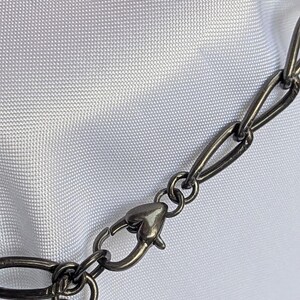 Puede incluir: Primer plano de una cadena de collar de metal gris oscuro con un cierre en forma de coraz&oacute;n. Los eslabones de la cadena son ovalados y est&aacute;n conectados por peque&ntilde;os anillos circulares. El fondo es una tela blanca texturizada.