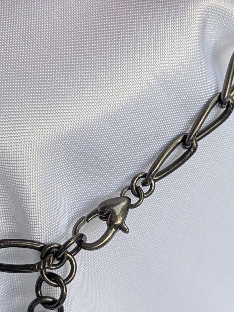 Puede incluir: Primer plano de una cadena de collar de metal gris oscuro con un cierre en forma de coraz&oacute;n. Los eslabones de la cadena son ovalados y est&aacute;n conectados por peque&ntilde;os anillos circulares. El fondo es una tela blanca texturizada.