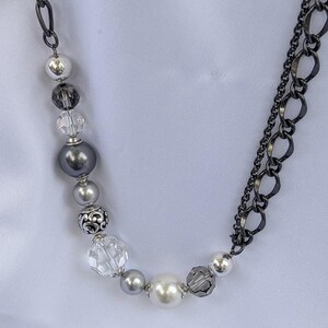Puede incluir: Un collar con una combinaci&oacute;n de cuentas plateadas, grises y de cristal transparente. Las cuentas incluyen perlas y cristales facetados, ensartados en una cadena oscura. El collar tiene un dise&ntilde;o en capas con una cadena en un lado y detalles de cuentas en el otro.