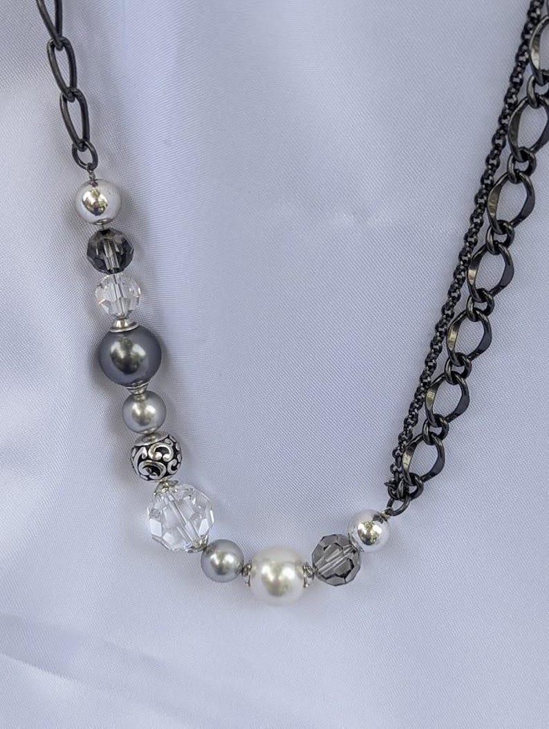 Puede incluir: Un collar con una combinaci&oacute;n de cuentas plateadas, grises y de cristal transparente. Las cuentas incluyen perlas y cristales facetados, ensartados en una cadena oscura. El collar tiene un dise&ntilde;o en capas con una cadena en un lado y detalles de cuentas en el otro.