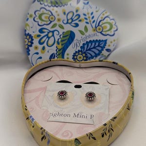 May include: A heart-shaped box with a floral pattern in blue, green, and yellow. Inside, silver stud earrings with red centres are presented on a card. The box is open, displaying the earrings and the brand name.