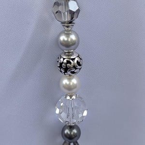 Puede incluir: Primer plano de un collar de cuentas con una variedad de cuentas plateadas y transparentes. Las cuentas incluyen cristales facetados, perlas y detalles decorativos plateados. El collar se muestra sobre un fondo gris claro.