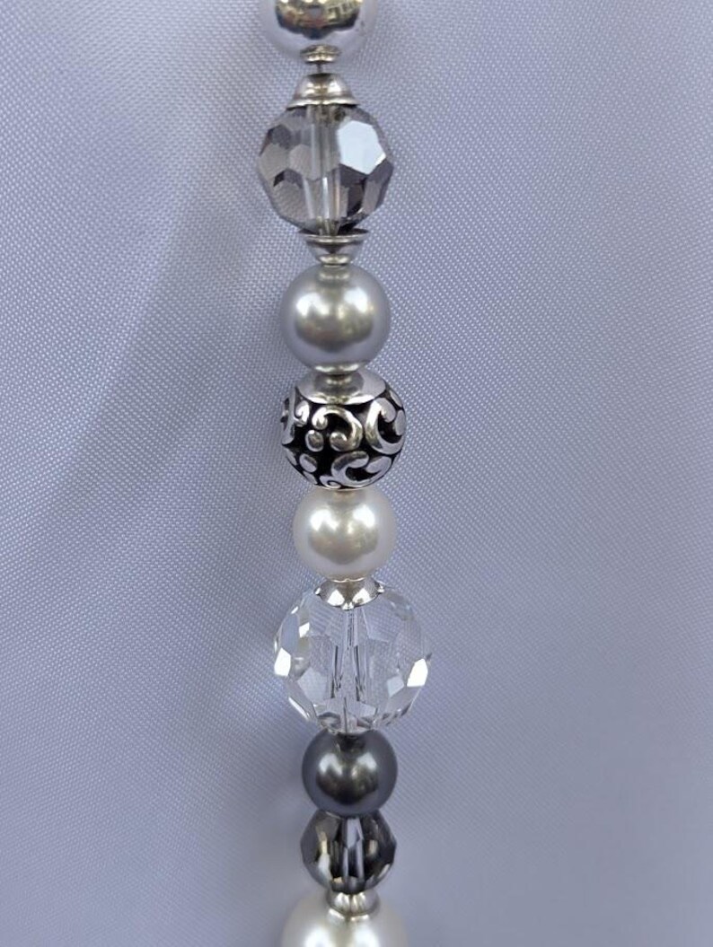 Puede incluir: Primer plano de un collar de cuentas con una variedad de cuentas plateadas y transparentes. Las cuentas incluyen cristales facetados, perlas y detalles decorativos plateados. El collar se muestra sobre un fondo gris claro.