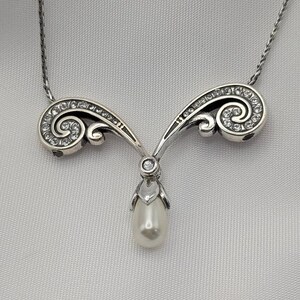 May include: A silver necklace with a pendant featuring a pear-shaped pearl. The pendant hangs from a decorative, swirling design set with small, clear gemstones. The necklace chain is delicate and silver.