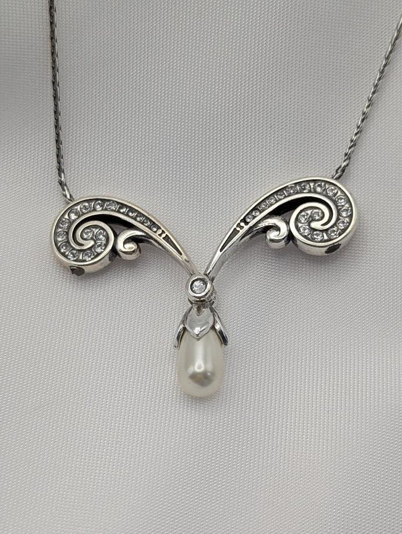 May include: A silver necklace with a pendant featuring a pear-shaped pearl. The pendant hangs from a decorative, swirling design set with small, clear gemstones. The necklace chain is delicate and silver.
