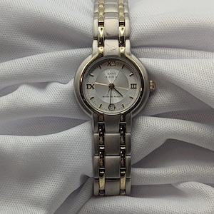 May include: Anne Klein watch with a silver and gold-tone metal band and a white face. The watch has Roman numeral hour markers and a date display at the 6 o'clock position. The watch is water resistant.