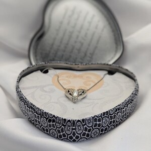 May include: A silver necklace with a heart-shaped pendant is displayed in a heart-shaped box. The box has a black and white decorative pattern. The necklace is set against a white backdrop.