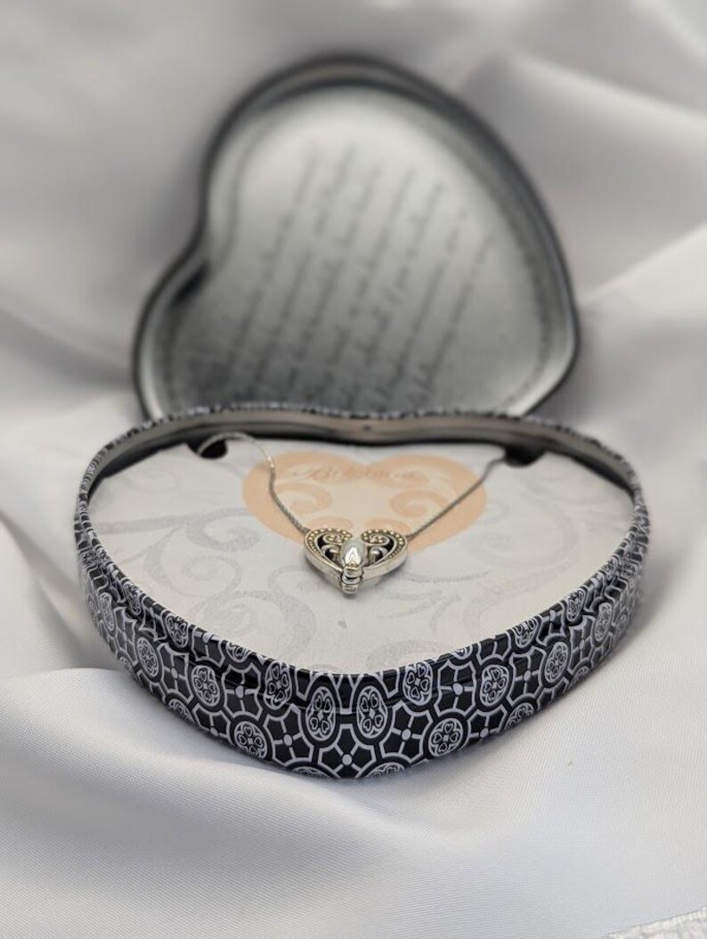 May include: A silver necklace with a heart-shaped pendant is displayed in a heart-shaped box. The box has a black and white decorative pattern. The necklace is set against a white backdrop.