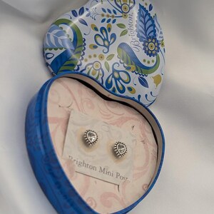 K&ouml;nnte beinhalten: Eine herzf&ouml;rmige blaue Schachtel mit floral gemustertem Deckel, die ein Paar Brighton Mini Post-Ohrringe enth&auml;lt. Die Ohrringe sind silberfarben mit einem klaren Stein in der Mitte. Die Schachtel ist ge&ouml;ffnet und zeigt die Ohrringe auf einer rosa Karte.