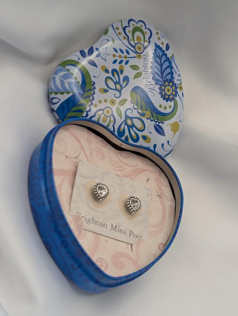 K&ouml;nnte beinhalten: Eine herzf&ouml;rmige blaue Schachtel mit floral gemustertem Deckel, die ein Paar Brighton Mini Post-Ohrringe enth&auml;lt. Die Ohrringe sind silberfarben mit einem klaren Stein in der Mitte. Die Schachtel ist ge&ouml;ffnet und zeigt die Ohrringe auf einer rosa Karte.