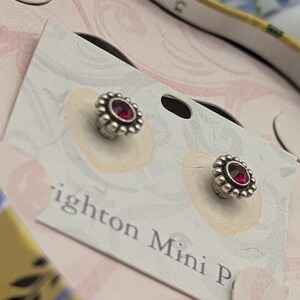 May include: Silver stud earrings, each featuring a central red gem encircled by a beaded border. The earrings are presented on a card with the text "Brighton Mini P". The background has a floral design.