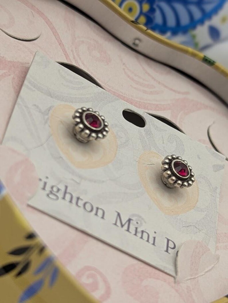 May include: Silver stud earrings, each featuring a central red gem encircled by a beaded border. The earrings are presented on a card with the text "Brighton Mini P". The background has a floral design.