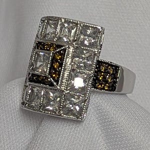 May include: A silver ring with a rectangular design featuring a cluster of clear and brown cubic zirconia stones.
