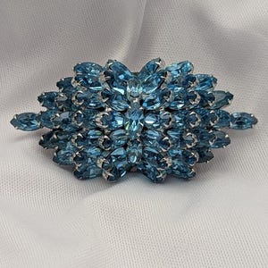 May include: A vintage brooch featuring a cluster of light blue, marquise-cut rhinestones. The stones are set in a silver-toned metal frame, creating a floral or starburst design. The brooch is a classic piece of jewelry.