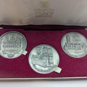 May include: A set of three circular silver-toned metal medallions housed in a maroon velvet-lined presentation case. The medallions feature embossed architectural illustrations of St. Paul's Cathedral, Westminster Abbey, and Westminster Cathedral.