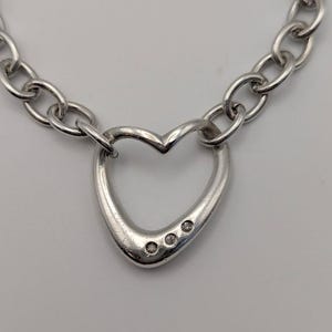 May include: A silver-toned chain necklace featuring a central open heart-shaped pendant. The bottom curve of the heart is accented with three small, clear, round-cut stones embedded in the metal surface, connected to a thick, polished oval link chain.