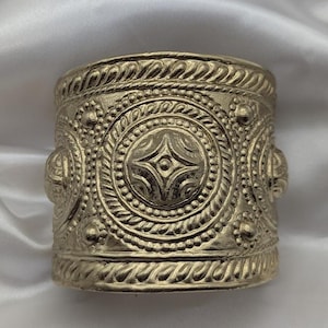 May include: A wide gold-toned cuff bracelet featuring an intricate embossed pattern. The design includes a central circular motif with a four-pointed star shape, surrounded by beaded and twisted rope textures, with additional circular side accents.