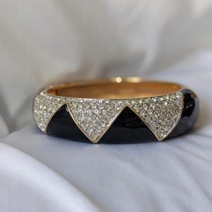 May include: A black and gold bangle bracelet with a row of clear rhinestones around the edge. The bracelet has a geometric design with three black triangles.