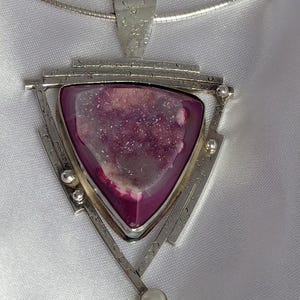May include: A silver pendant necklace featuring a triangular, magenta-colored stone with a pearl accent. The pendant has a geometric design with silver bars and a small pearl at the bottom. The necklace is on a silver chain.