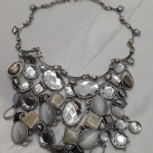 May include: A statement necklace featuring a dark silver-toned metal chain adorned with an array of faceted clear crystals, smooth grey cabochons, and rectangular beige stones. The design includes multiple dangling elements in various shapes.