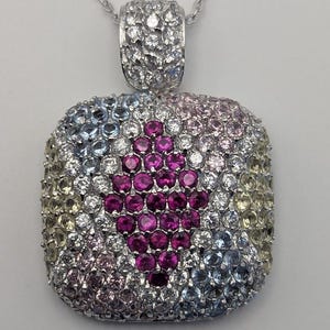 May include: A silver pendant necklace featuring a square pendant covered in small, colorful gemstones. The pendant has a central design of deep pink gems, surrounded by a mix of blue, pink, yellow, and clear stones. The necklace chain is also silver.