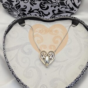 May include: A silver heart pendant necklace with a pearl-like centre, presented in a heart-shaped box. The box features a black and white floral pattern on the lid and a cream interior with the word "Brighton".