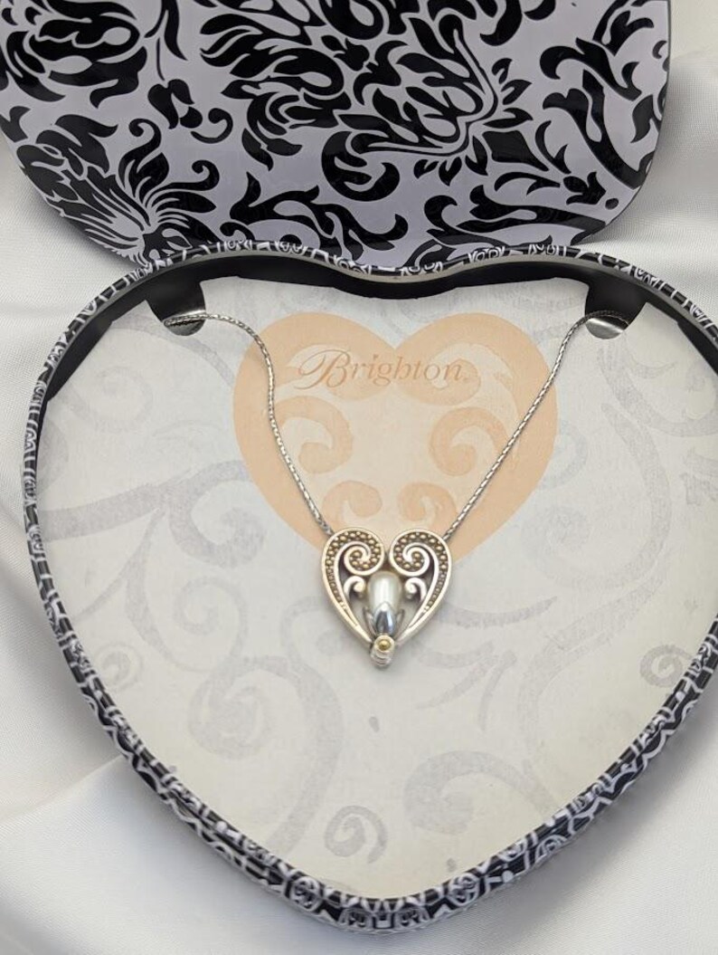 May include: A silver heart pendant necklace with a pearl-like centre, presented in a heart-shaped box. The box features a black and white floral pattern on the lid and a cream interior with the word "Brighton".