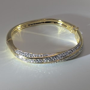 May include: A gold-toned hinged bangle bracelet. The bracelet features two rows of small, clear crystals set along the top half of the band. The bracelet is on a white surface.