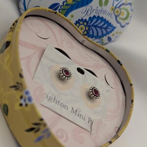 May include: Silver stud earrings with a small, round, red gemstone centre, displayed in a heart-shaped tin. The tin is yellow with a floral design and the brand name "Brighton" is visible.