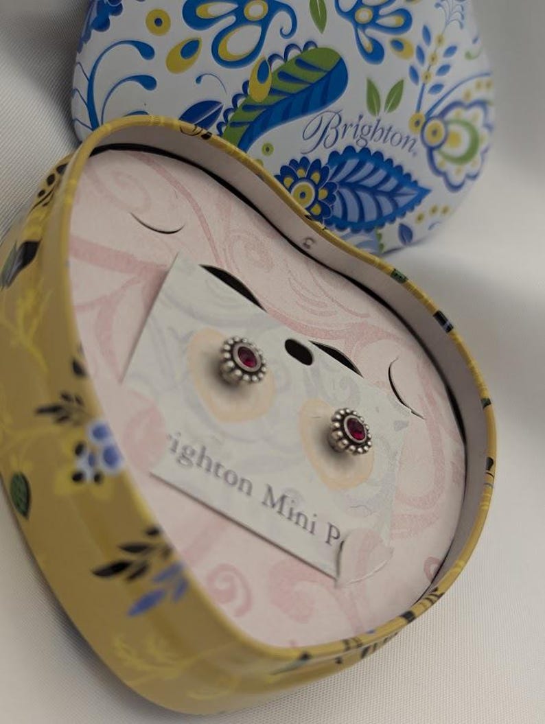 May include: Silver stud earrings with a small, round, red gemstone centre, displayed in a heart-shaped tin. The tin is yellow with a floral design and the brand name "Brighton" is visible.