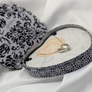 May include: A heart-shaped jewellery box with a black and white floral pattern, open to reveal a silver necklace with a heart-shaped pendant. The box is on a white fabric background.