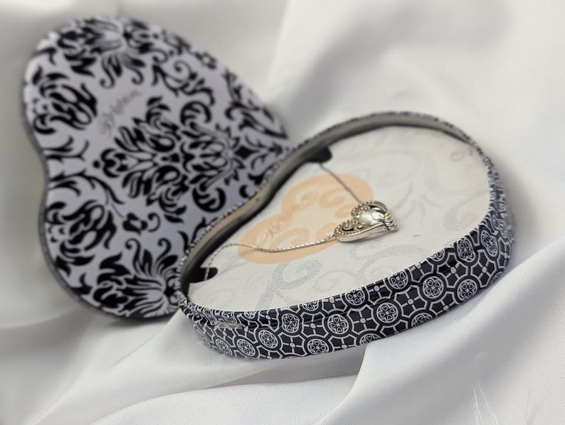 May include: A heart-shaped jewellery box with a black and white floral pattern, open to reveal a silver necklace with a heart-shaped pendant. The box is on a white fabric background.