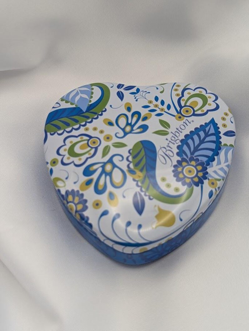 K&ouml;nnte beinhalten: Herzf&ouml;rmige dekorative Dose mit blauem, gr&uuml;nem und gelbem Blumenmuster auf wei&szlig;em Hintergrund. Die Dose hat einen blauen Rand und das Wort "Brighton" ist auf dem Deckel aufgedruckt.