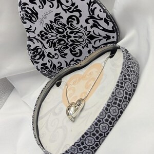 May include: A heart-shaped jewellery box with a black and white floral design, opened to reveal a silver necklace with a heart pendant. The box is on a white fabric surface. The brand name "Brighton" is visible.