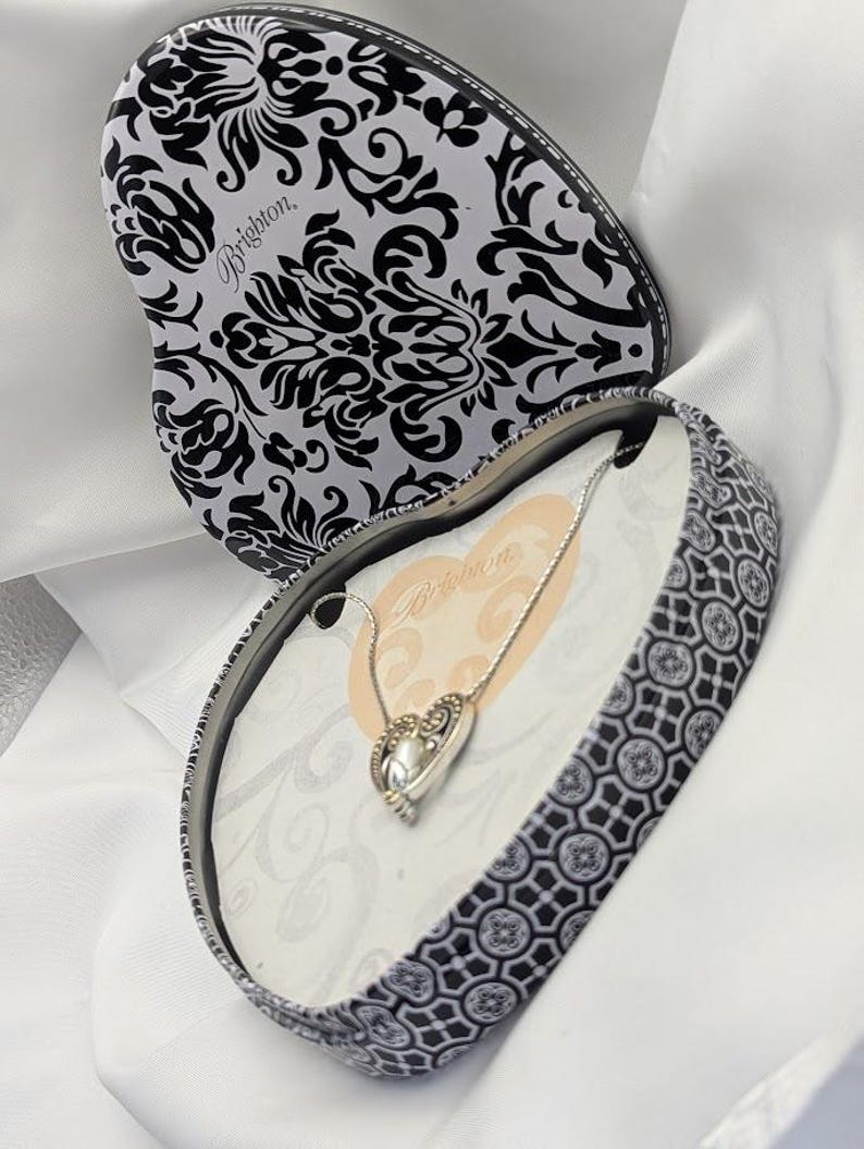 May include: A heart-shaped jewellery box with a black and white floral design, opened to reveal a silver necklace with a heart pendant. The box is on a white fabric surface. The brand name "Brighton" is visible.