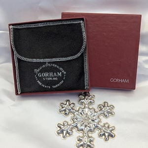 May include: A silver snowflake ornament with a detailed design, resting on a white surface. The ornament is next to a red box and a black pouch with the text "PACIFIC SILVER CLOTH" and "GORHAM STERLING" printed on it.