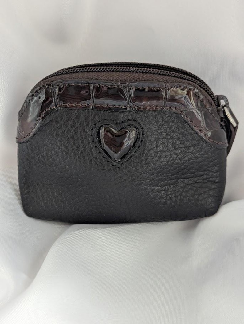 以下が含まれることがあります： ハート型のアクセントが付いた、小さな濃い茶色の小銭入れ。 質感のある革のボディと、光沢のあるクロコダイルエンボス加工のトップ部分が特徴です。 ジッパー開閉と小さなプルが付いています。