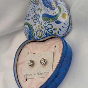 K&ouml;nnte beinhalten: Eine herzf&ouml;rmige blaue Dose mit floralem Design, ge&ouml;ffnet, um ein Paar herzf&ouml;rmiger Ohrstecker auf einer Karte zu enth&uuml;llen. Die Ohrringe haben einen silberfarbenen, perlenbesetzten Rand und einen klaren Mittelstein. Der Deckel der Dose ist mit blauen, gr&uuml;nen und wei&szlig;en Blumenmustern verziert.