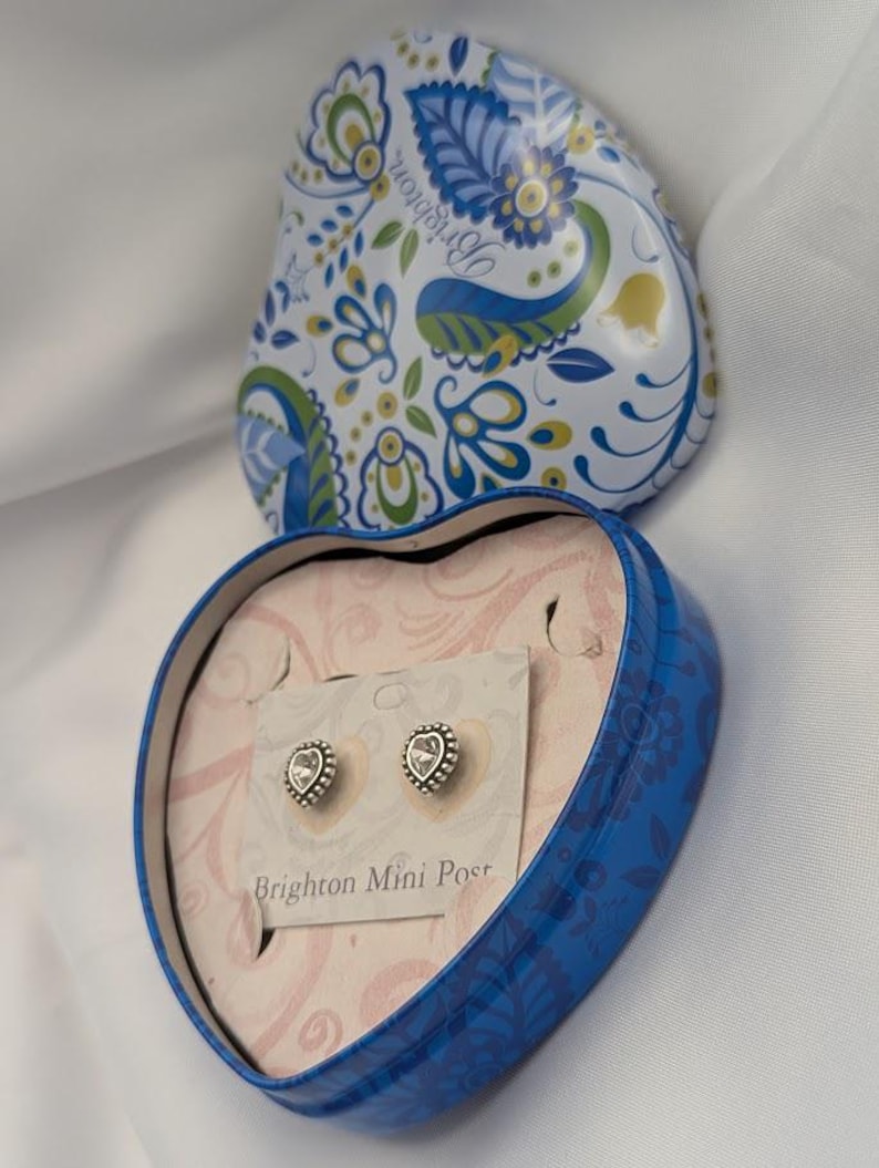 K&ouml;nnte beinhalten: Eine herzf&ouml;rmige blaue Dose mit floralem Design, ge&ouml;ffnet, um ein Paar herzf&ouml;rmiger Ohrstecker auf einer Karte zu enth&uuml;llen. Die Ohrringe haben einen silberfarbenen, perlenbesetzten Rand und einen klaren Mittelstein. Der Deckel der Dose ist mit blauen, gr&uuml;nen und wei&szlig;en Blumenmustern verziert.