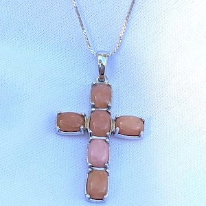 May include: A silver-toned necklace featuring a cross pendant. The cross is adorned with oval-shaped, peach-colored gemstones set in a silver frame. The necklace hangs on a delicate silver chain, set against a white background.
