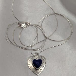 May include: A silver-toned necklace with a heart-shaped pendant. The pendant has a dark blue heart in the center, surrounded by silver with 'X' and 'O' symbols. The chain is a thin, delicate silver.