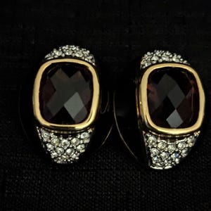 May include: A pair of gold tone clip-on earrings with a purple faceted stone set in a gold tone bezel. The stones are surrounded by a halo of clear rhinestones.