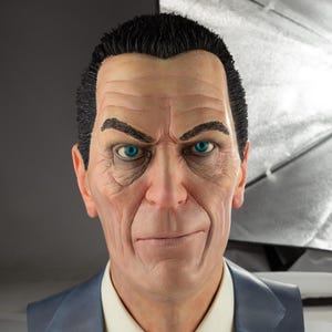 Puede incluir: Busto detallado de un hombre con pelo corto y negro, ojos azules intensos y expresión severa. Lleva una chaqueta de traje azul sobre una camisa blanca con cuello. La escultura tiene tonos de piel y detalles faciales realistas.