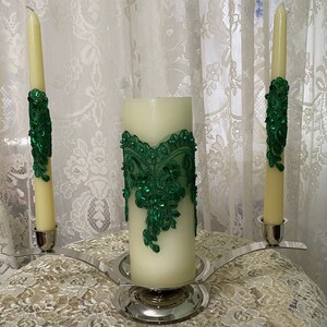 May include: A silver candle holder with three candles. The center candle is a large pillar candle with a green lace design. The two smaller candles are on either side of the center candle and have green lace designs around the bottom.