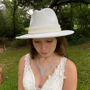 White bridal wedding fedora hat with hat band of rows of ivory pears. The back is accented with a white ribbon bow.