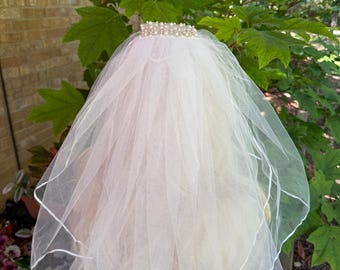 May include: A white bridal veil with a pearl-adorned tiara. The veil is made of sheer fabric and has a delicate, layered design. The edges are trimmed with a thin white ribbon. The background features green foliage and a brick wall.