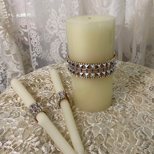 Puede incluir: Dos velas de té blancas con bandas decorativas plateadas y doradas, y una vela pilar blanca más grande con una banda similar. Las velas están sobre un mantel de encaje blanco.