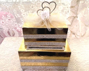 Caja de cartón de oro metálico de dos niveles, adornada con filas de perlas y diamantes de imitación, con una rosa blanca y dos corazones dorados decorados con diamantes de imitación.
