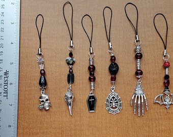 May include: Seven black and silver charm phone charms with a gothic theme. The charms include a skull, a raven, a coffin, a spider web, a skeleton hand, a bat, and a crystal bead.